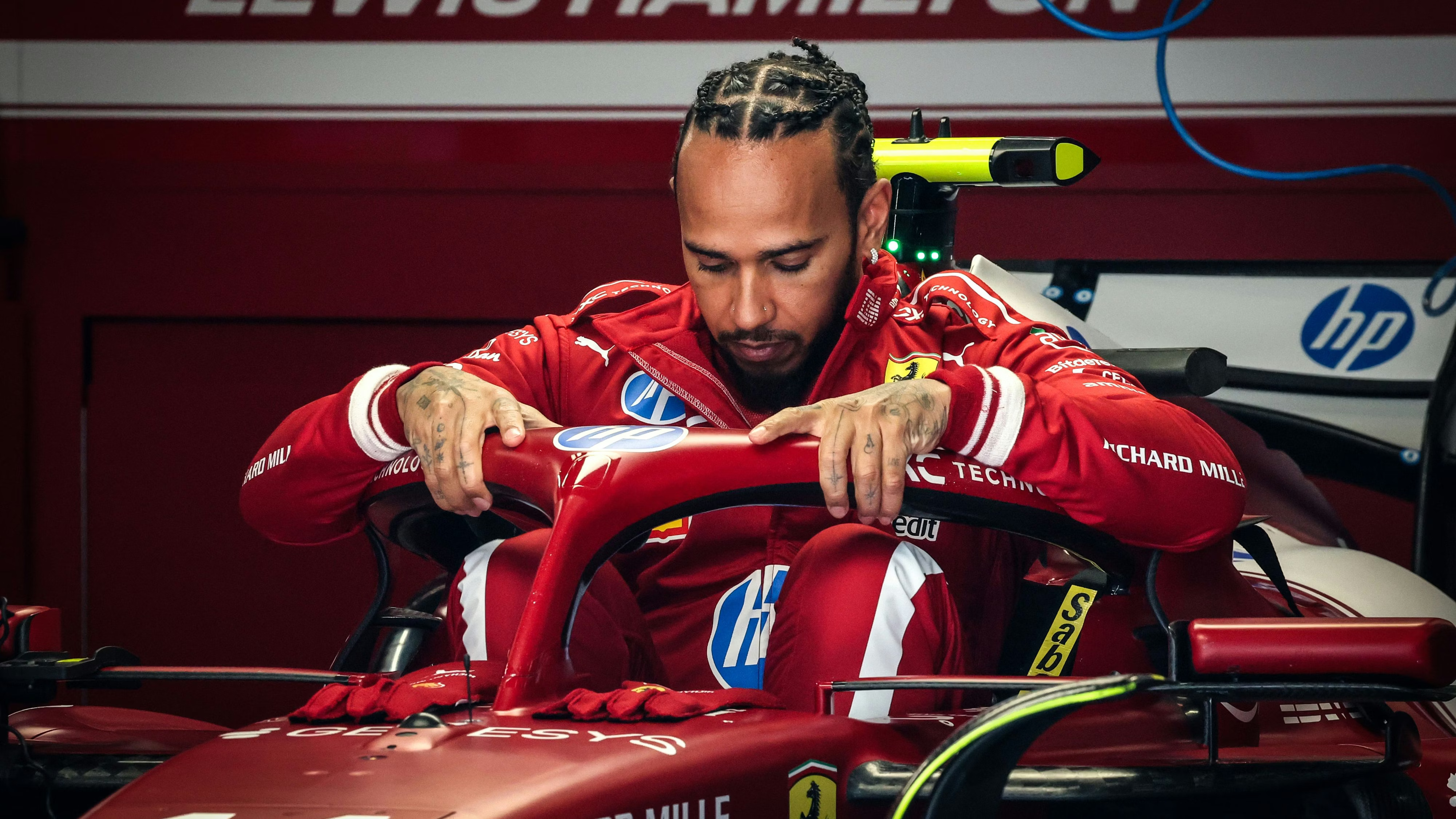 Lewis Hamilton getting into a red Ferrari Formula 1 car. You can see the Halo of the car and a red backdrop