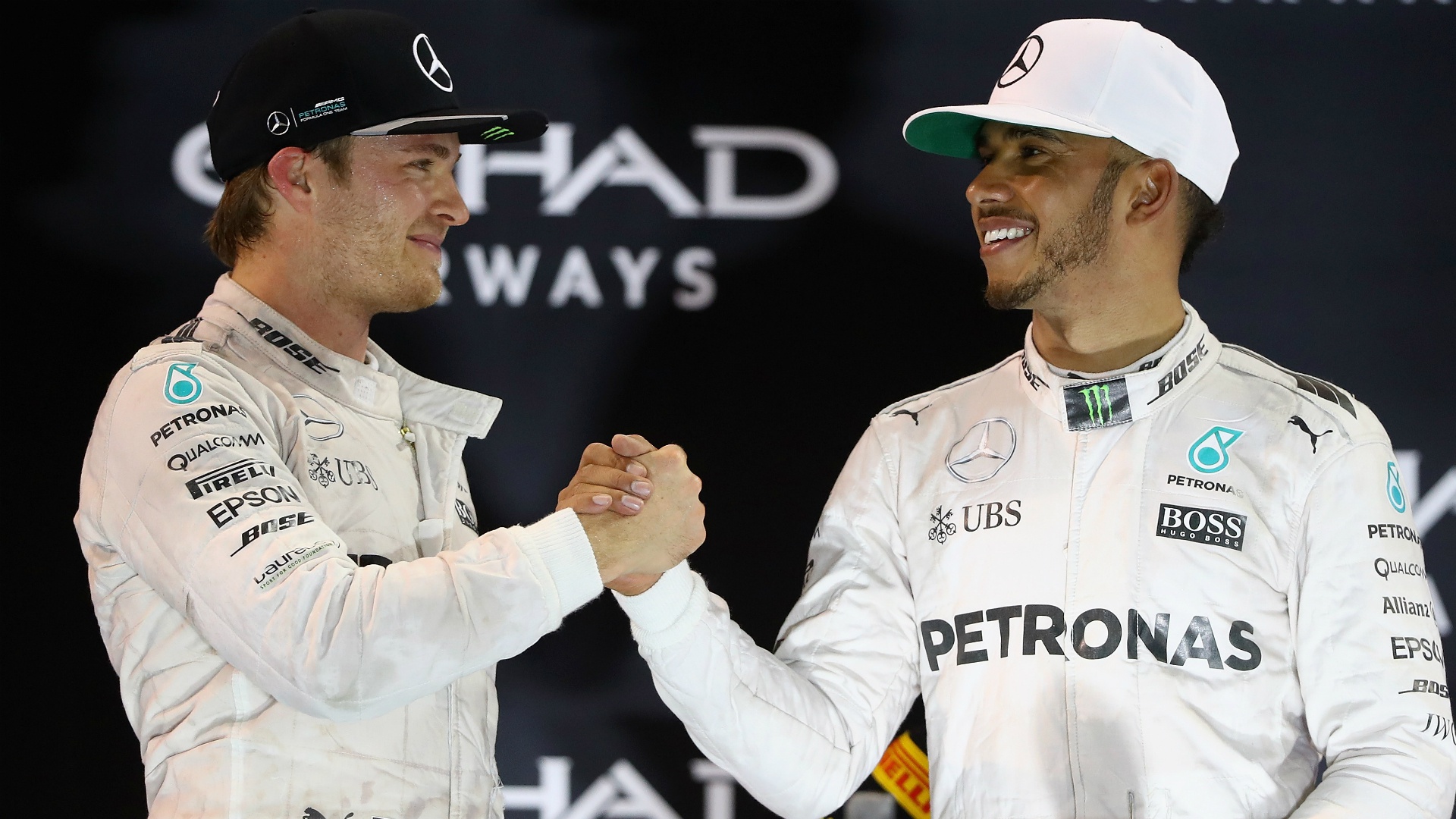 Nico Rosberg and Lewis Hamilton clasp hands. Both are wearing white racing suits with Hamilton wearing a white ballcap and Rosberg wearing a black one. They stand in front of a dark Etihad Airways background